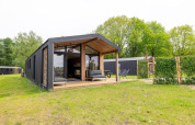 Lodge moderne avec grande terrasse et façade vitrée, entouré de pelouse verte et d’arbres dans un cadre naturel.