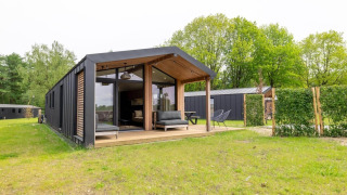 Lodge moderne avec grande terrasse et façade vitrée, entouré de pelouse verte et d’arbres dans un cadre naturel.