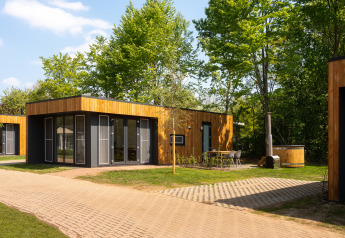 Moderne houten lodge met grote ramen, buitenterras en een hottub omringd door groen en bomen.