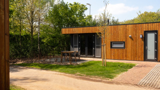 Außenansicht einer modernen Holzhütte mit Terrasse, Gartenmöbeln und viel Grün in einer Lodge.