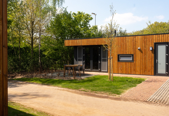 Vista exterior de una moderna cabaña de madera con terraza, muebles de jardín y rodeada de naturaleza.