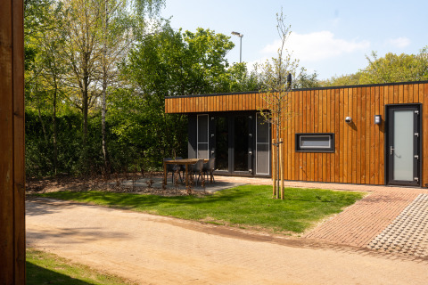 Außenansicht einer modernen Holzhütte mit Terrasse, Gartenmöbeln und viel Grün in einer Lodge.