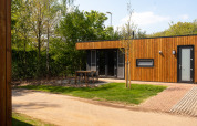 Outdoor view of a modern wooden lodge with patio, outdoor furniture, green grass, and trees around.
