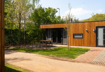 Außenansicht einer modernen Holzhütte mit Terrasse, Gartenmöbeln und viel Grün in einer Lodge.