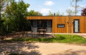 A modern wood-paneled lodge with a patio and garden, surrounded by trees under a clear blue sky.