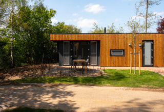 Een moderne houten lodge met terras en tuin, omringd door bomen onder een heldere blauwe lucht.