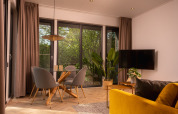 Stijlvolle lodge woonkamer met grote ramen, glazen tafel, grijze stoelen, gele bank en uitzicht op de natuur.