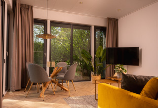 Acogedora sala de lodge con ventanas grandes, mesa de cristal, sillas grises, sofá amarillo y plantas.