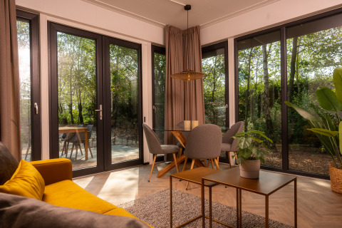 Lichte, moderne lodge-living met gele zetel, eettafel, planten en grote ramen met zicht op het bos.