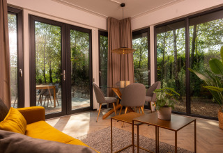 Lyst og moderne lodge-stue med gul sofa, spisebord, grønne planter og store vinduer med udsigt til skov.