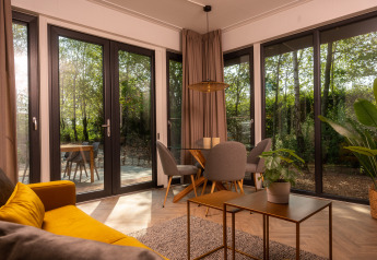 Luminoso salón moderno de lodge con sofá amarillo, comedor, plantas y grandes ventanales con vista al bosque.