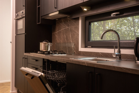 Cocina moderna en una cabaña con estufa de gas, fregadero, lavavajillas y ventana con vistas al bosque.