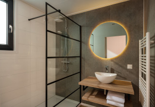 Moderne lodgebadkamer met inloopdouche, verlicht rond spiegel, houten wastafel en handdoeken.