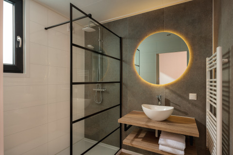 Bagno moderno di un lodge con doccia a pioggia, specchio rotondo illuminato e lavabo su piano in legno.