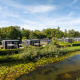 Flotte tiny houses ved en flod med åkander, omgivet af grønt, under en blå himmel, kaldet River Cabin.