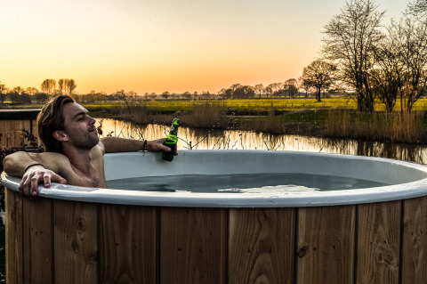 Hombre relajándose en un jacuzzi con cerveza junto al río al atardecer en Holiday Park Mölke, Países Bajos.