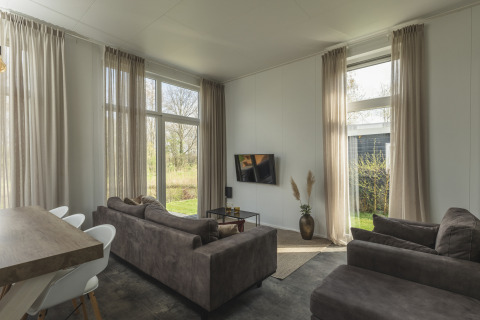 Wohnzimmer der River Cabin mit großen Fenstern, braunen Sofas und TV im Holiday Park Mölke in den Niederlanden.