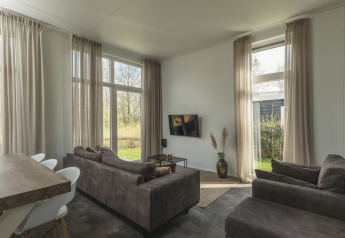Woonkamer van River Cabin met grote ramen, bruine banken, tv en natuurzicht bij Holiday Park Mölke, Nederland.