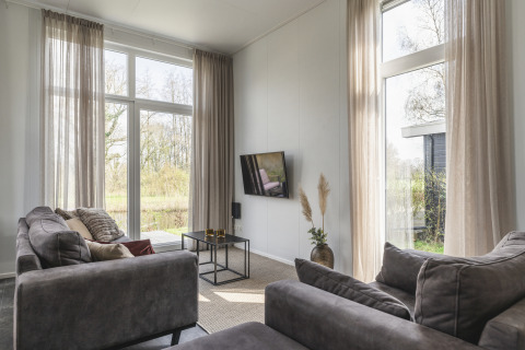 Woonkamer in River Cabin bij Holiday Park Mölke, Nederland, met grote ramen, grijze banken en uitzicht op natuur.