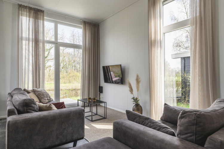 Salón en River Cabin del Holiday Park Mölke, Países Bajos, con ventanales grandes, sofás grises y vista natural.