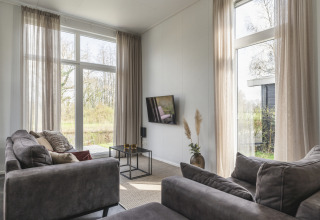 Woonkamer van River Cabin in Holiday Park Mölke, Nederland, met grote ramen en grijze zetels met natuurzicht.