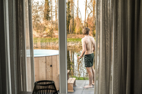 Man in shorts stands by a terrace overlooking a lake at River Cabin + hot tub at Holiday Park Mölke, Netherlands.