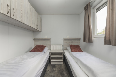 Moderne glampingkamer met twee eenpersoonsbedden, kussens in bruintint, opbergkasten en een raam met gordijnen.