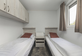 Moderne glampingkamer met twee eenpersoonsbedden, kussens in bruintint, opbergkasten en een raam met gordijnen.