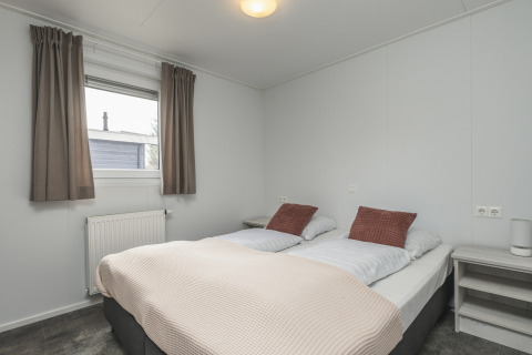 Lichte glamping slaapkamer met tweepersoonsbed, nachtkastjes, raam met gordijnen en radiator.
