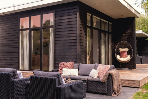 Terraza moderna exterior junto a una casa pequeña de madera negra con sillones y silla colgante en Holanda.