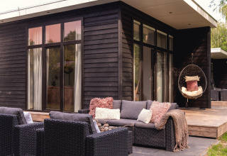 Terraza moderna exterior junto a una casa pequeña de madera negra con sillones y silla colgante en Holanda.