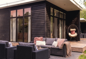 Terraza moderna exterior junto a una casa pequeña de madera negra con sillones y silla colgante en Holanda.