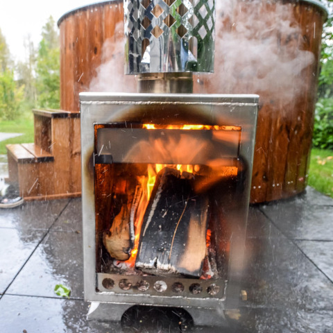 Poêle à bois extérieur avec feu allumé devant un bain à remous fumant à River Cabin, Holiday Park Mölke.