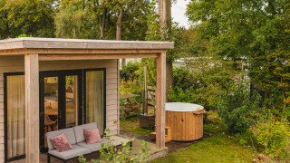 Gemütliches River Lodge Tiny House mit Terrasse, Sofa und Hot Tub, umgeben von grüner Natur.
