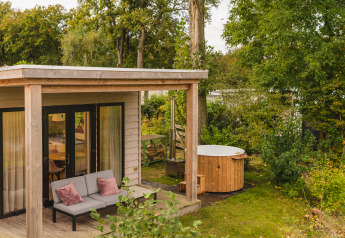 Gemütliches River Lodge Tiny House mit Terrasse, Sofa und Hot Tub, umgeben von grüner Natur.