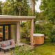 Hyggelig River Lodge tiny house med terrasse, sofa og udendørs træ-hot tub omgivet af grøn natur.