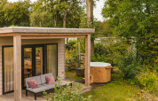 Hyggelig River Lodge tiny house med terrasse, sofa og udendørs træ-hot tub omgivet af grøn natur.