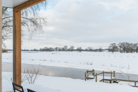 Blick von der Terrasse auf verschneite Felder und Fluss bei River lodge + hot tub, Mölke, Niederlande.