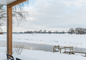 Blick von der Terrasse auf verschneite Felder und Fluss bei River lodge + hot tub, Mölke, Niederlande.