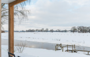View from patio of River lodge + hot tub at Holiday Park Mölke, snowy river and field in the Netherlands.