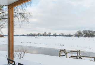 View from patio of River lodge + hot tub at Holiday Park Mölke, snowy river and field in the Netherlands.
