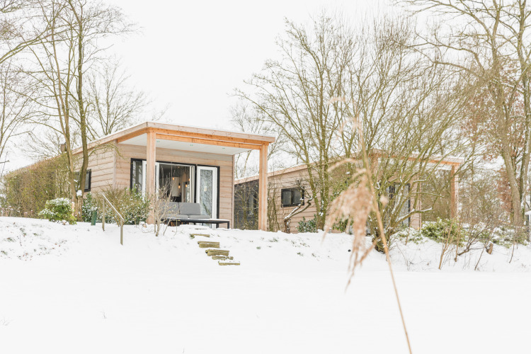 Pequeña casa con porche de madera y nieve en Holiday Park Mölke, Países Bajos, rodeada de árboles.