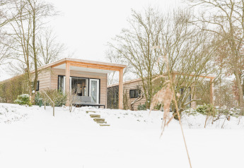 Petite maison avec terrasse en bois dans la neige au Holiday Park Mölke aux Pays-Bas, entourée d’arbres.