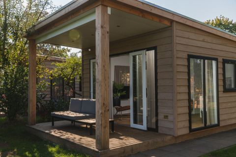 Modern glamping cabin with wood paneling, covered porch, and outdoor seating set in a lush, green area.