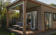 Alloggio glamping moderno in legno con veranda coperta e divano esterno, immerso nel verde.