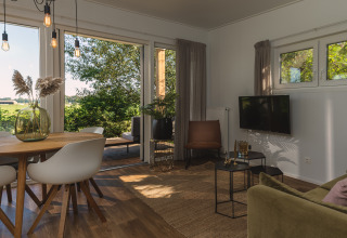 Lichte en moderne glamping woonkamer met eettafel, stoelen en grote ramen met uitzicht op de natuur.