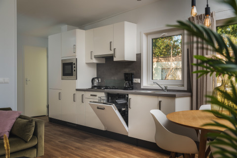 Moderne glampingkeuken met open vaatwasser, eettafel, groene zetel en groot raam met uitzicht.