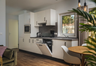 Moderne glampingkeuken met open vaatwasser, eettafel, groene bank en groot raam met uitzicht.