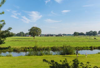 Veduta di una glamping: prati verdi, mucche, un ruscello e una famiglia che passeggia sul prato.