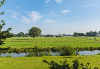 Veduta di una glamping: prati verdi, mucche, un ruscello e una famiglia che passeggia sul prato.